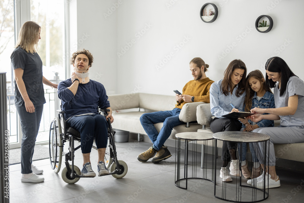 Young people in the lobby of the clinic. Rehabilitologist or nurse taking care of patient in wheelch