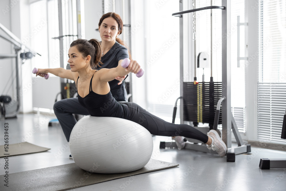 Woman doing exercises on fitness ball and dumbbells with rehabilitation specialist at the gym. Conce