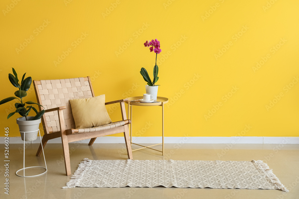 Orchid flower, cup of coffee on table and armchair near yellow wall in room