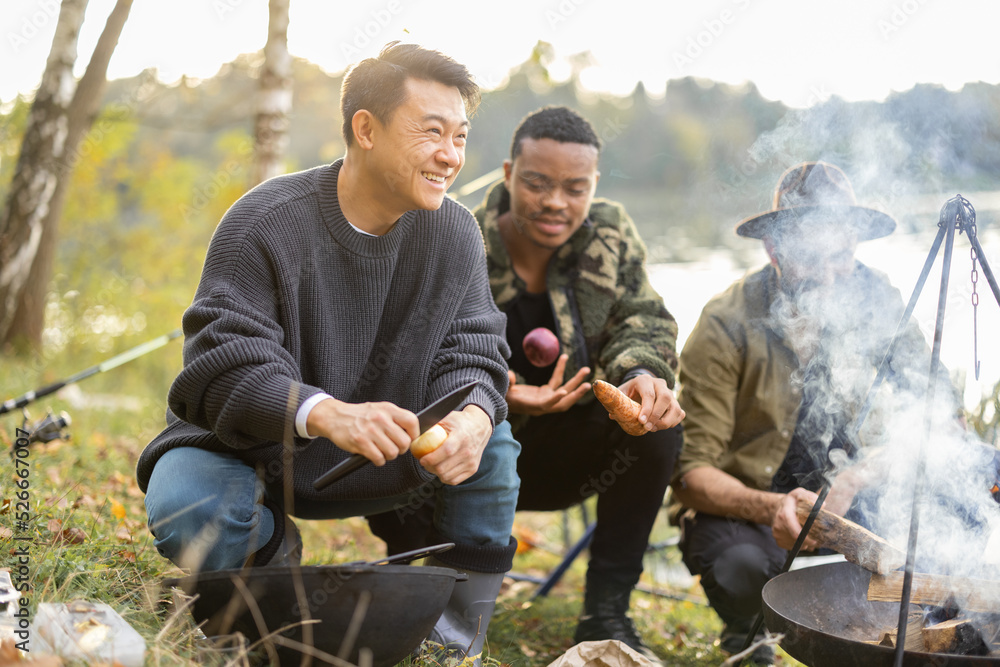 三个多种族的朋友在野餐时用篝火在大锅里煮乌卡汤。男人们在钓鱼和