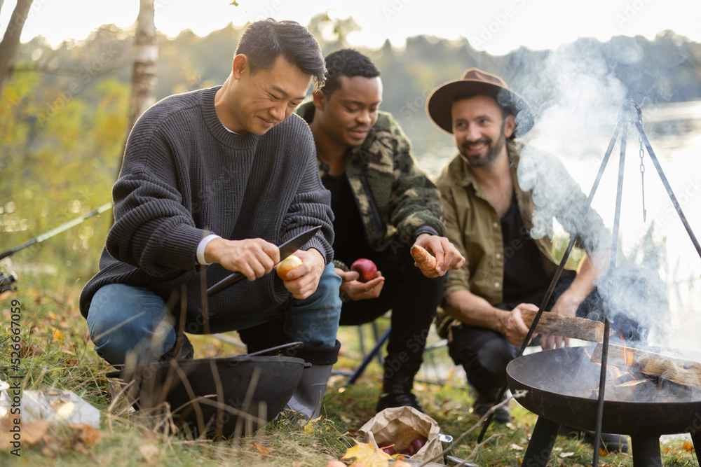 三个多种族的朋友在野餐时用篝火在大锅里煮乌卡汤。男人们在钓鱼和