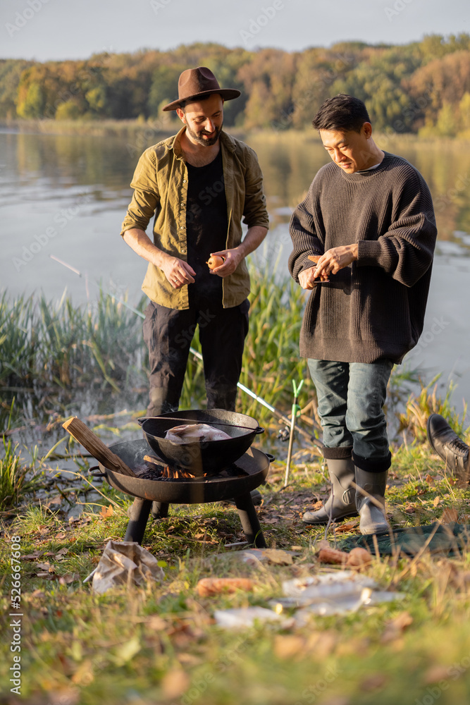 Two multiracial friends cooking Ukha soup in cauldron on bonfire during a picnic. Men talking and ha