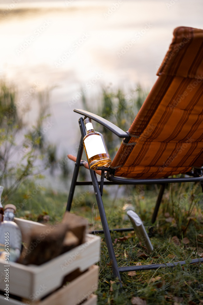 Bottle with strong alcohol and flask on folding chair at picnic or fishing. Bottle with blank label 