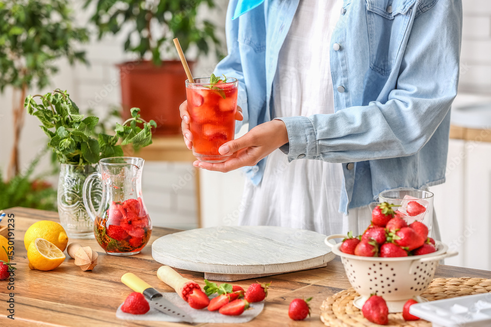 Woman holding glass of cold strawberry lemonade in kitchen