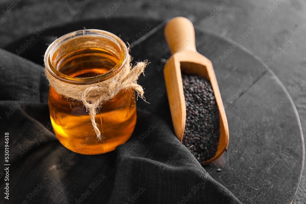 Board with jar of oil and sesame seeds on dark background, closeup