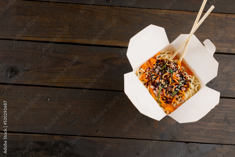 Overhead view of take away asian wok stir fry noodles with chopsticks in paper container on wood