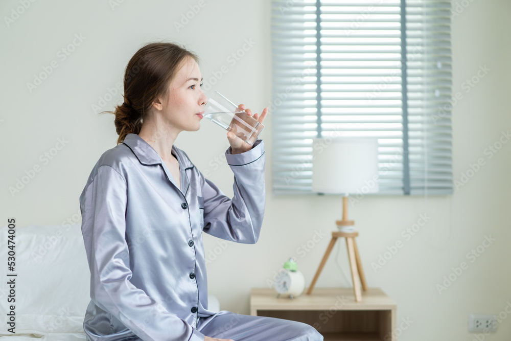 Young Caucasian woman in pajamas drink a glass of water in kitchen.