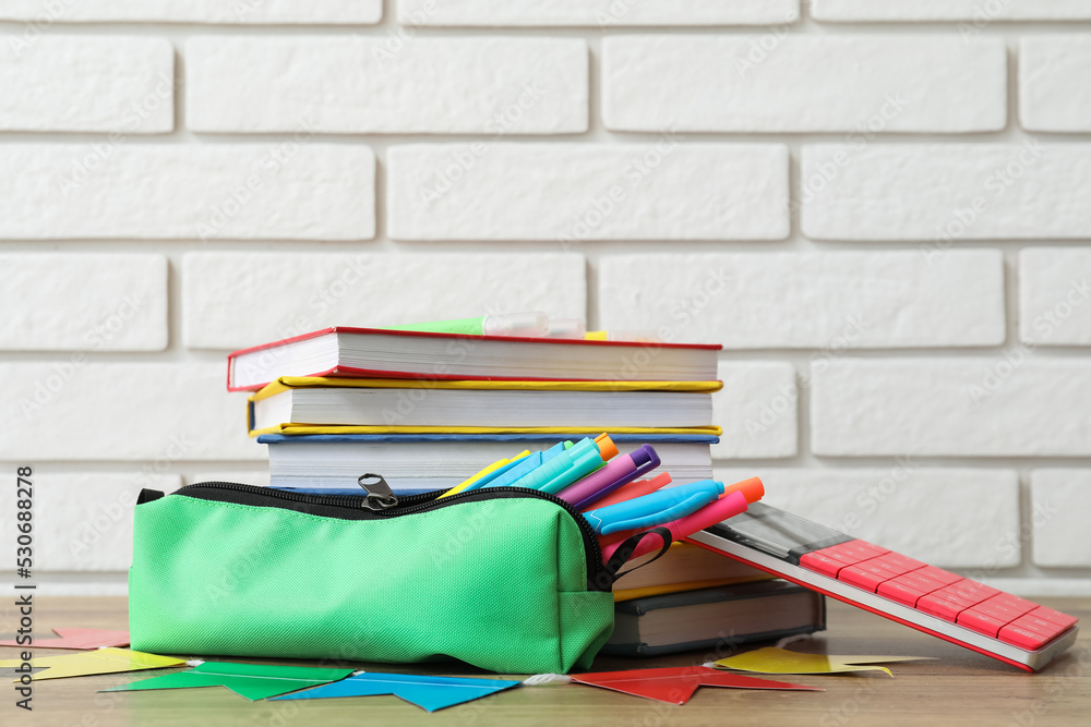 Green pencil case with school stationery and books on table near white brick wall