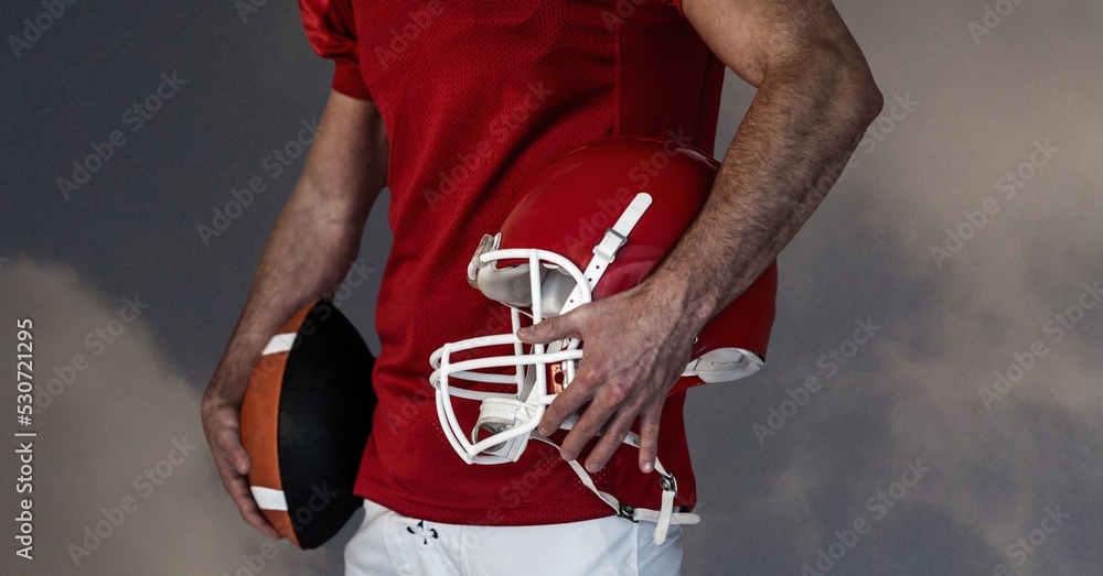 Composition of male american football player holding ball and helmet over blue sky with clouds