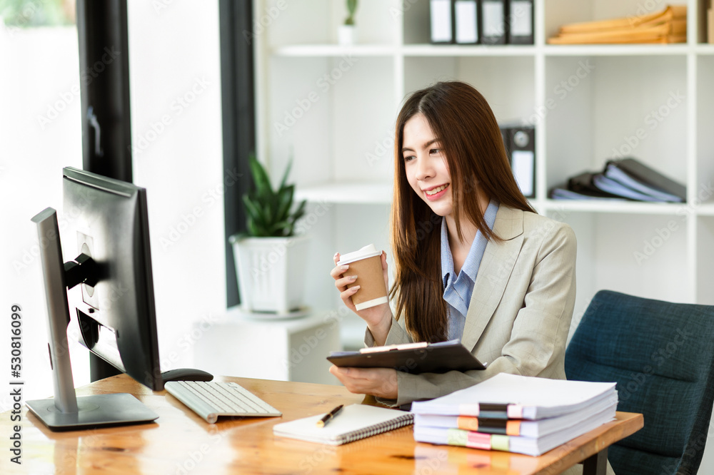 Confident beautiful Asian businesswoman typing laptop computer and digital tablet while holding coff