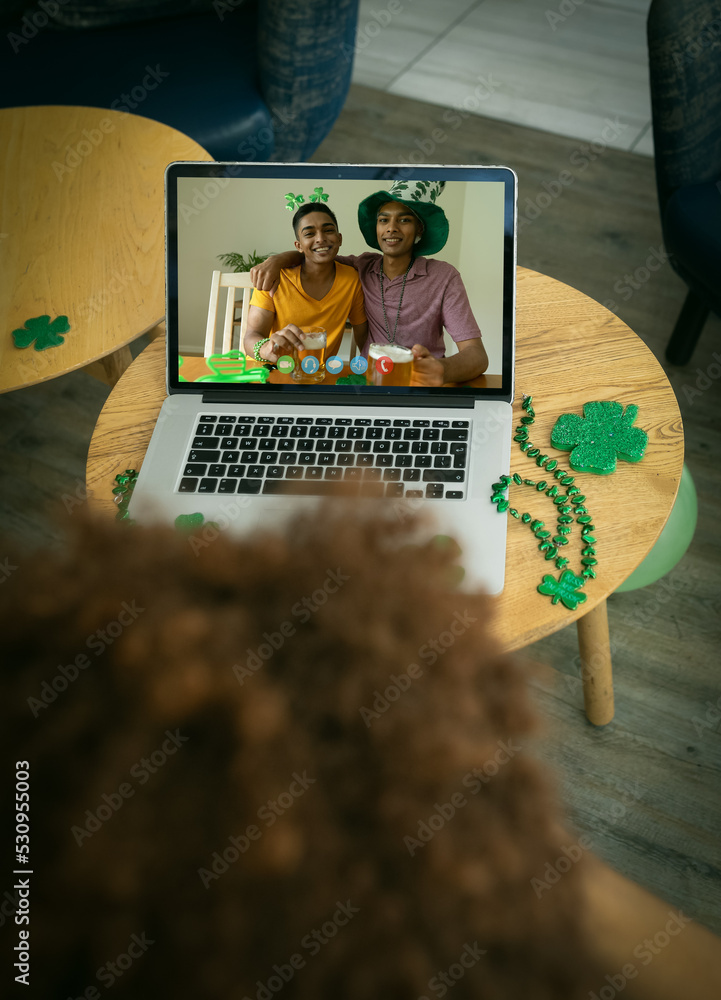 Mixed race man at bar making st patricks day video call to male friends embracing on laptop screen