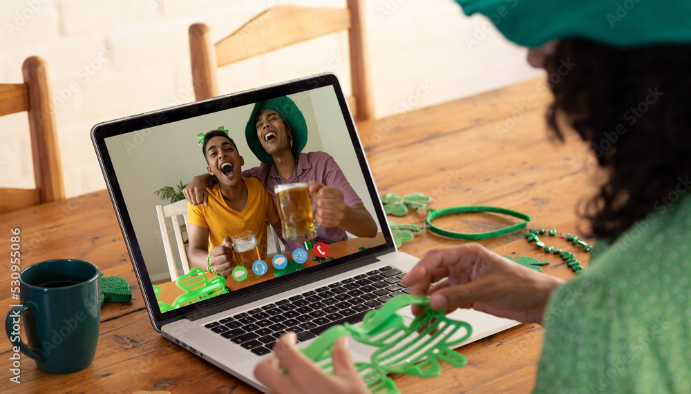 Mixed race woman making st patricks day video call to male friends drinking beer on laptop at home