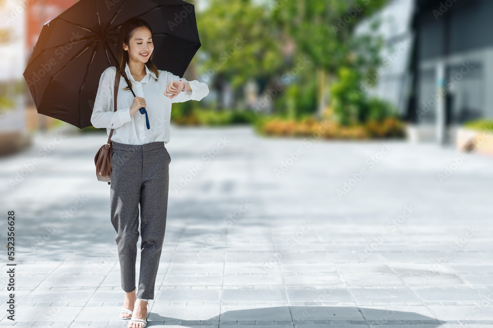 阳光明媚的日子里，带着黑色雨伞在户外看手表的可爱亚洲商业女性，专注