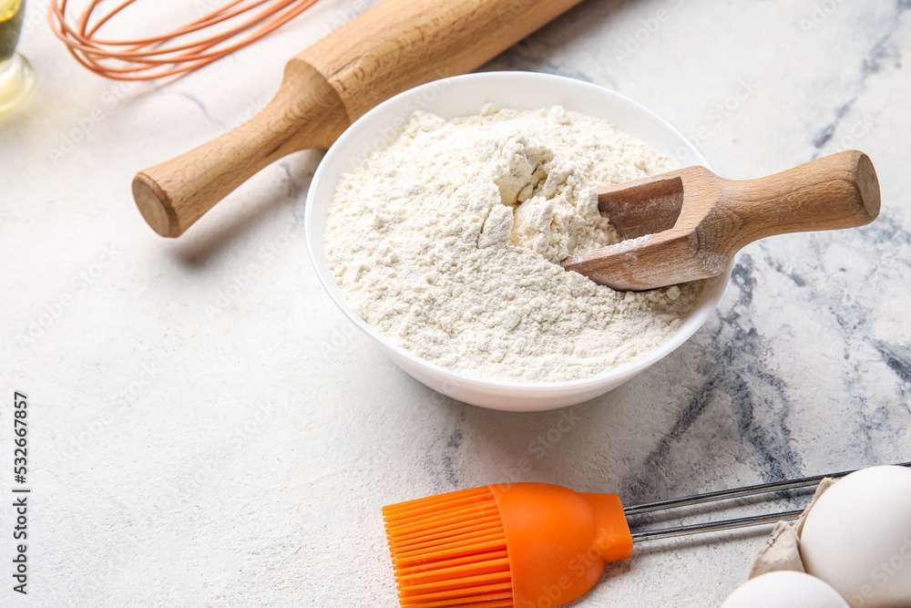 Bowl with flour, eggs and kitchen utensils on light table, closeup