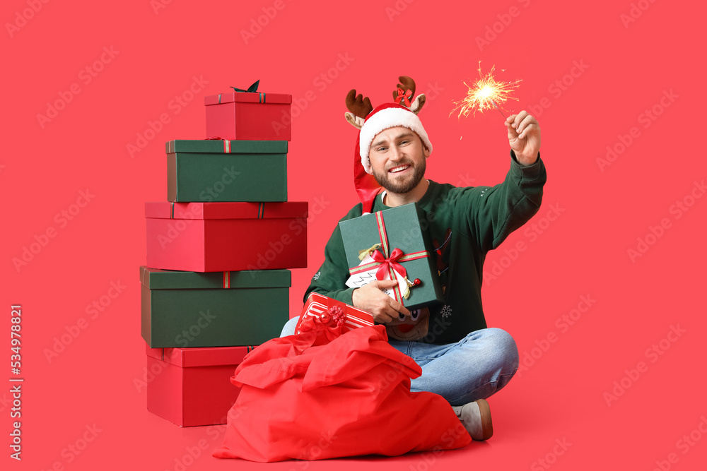 Happy man with Christmas gifts, sparkler and Santa bag on red background