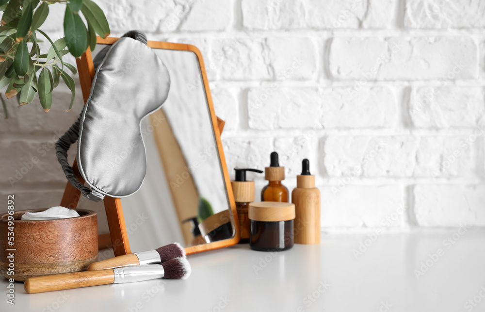 Mirror with sleeping mask, cosmetics and makeup brushes on table near white brick wall