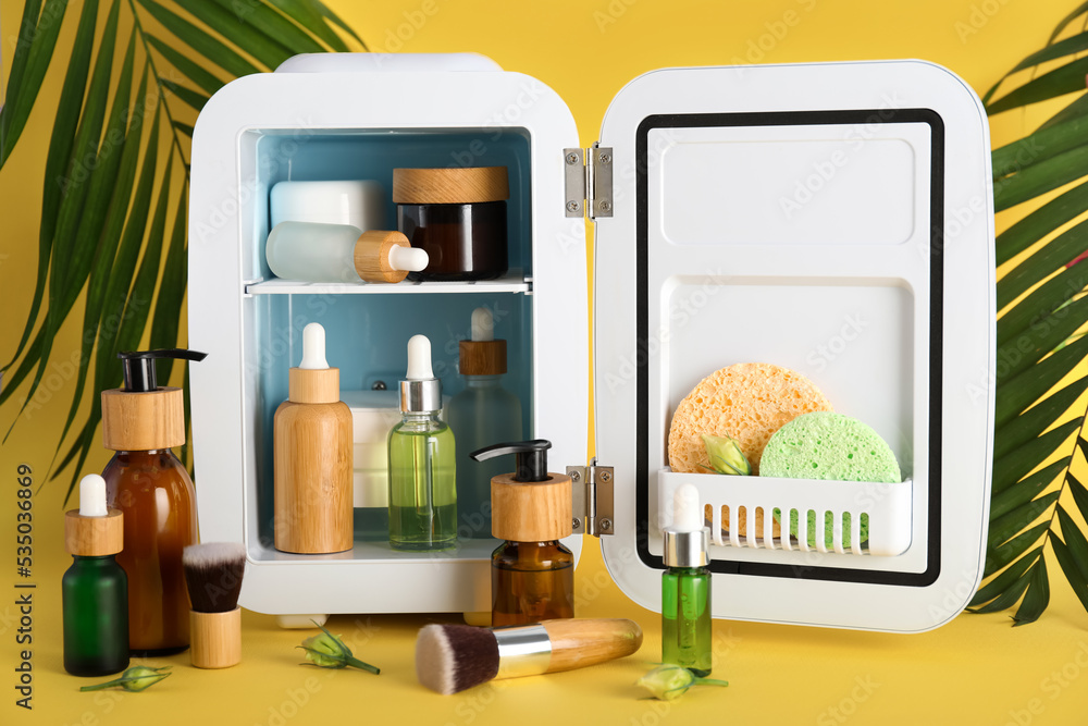 Small refrigerator with cosmetics and tropical palm leaves on yellow background