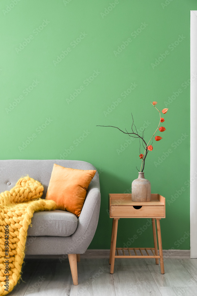 Vase with tree branch on table and grey sofa near green wall