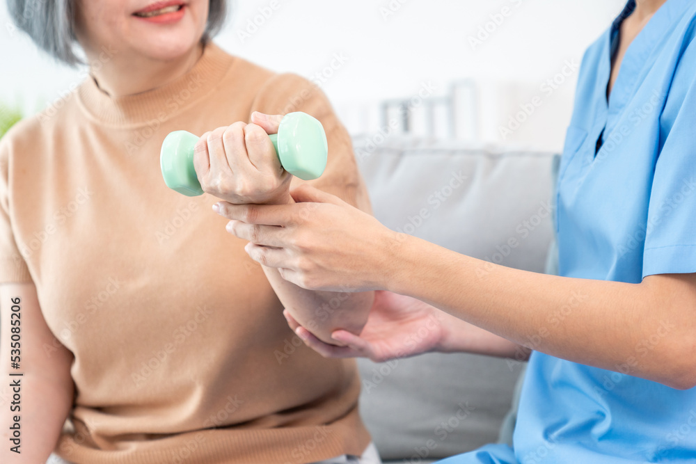 Caregiver helping contented senior woman exercise with dumbbell at home. Rehab for elder, senior med