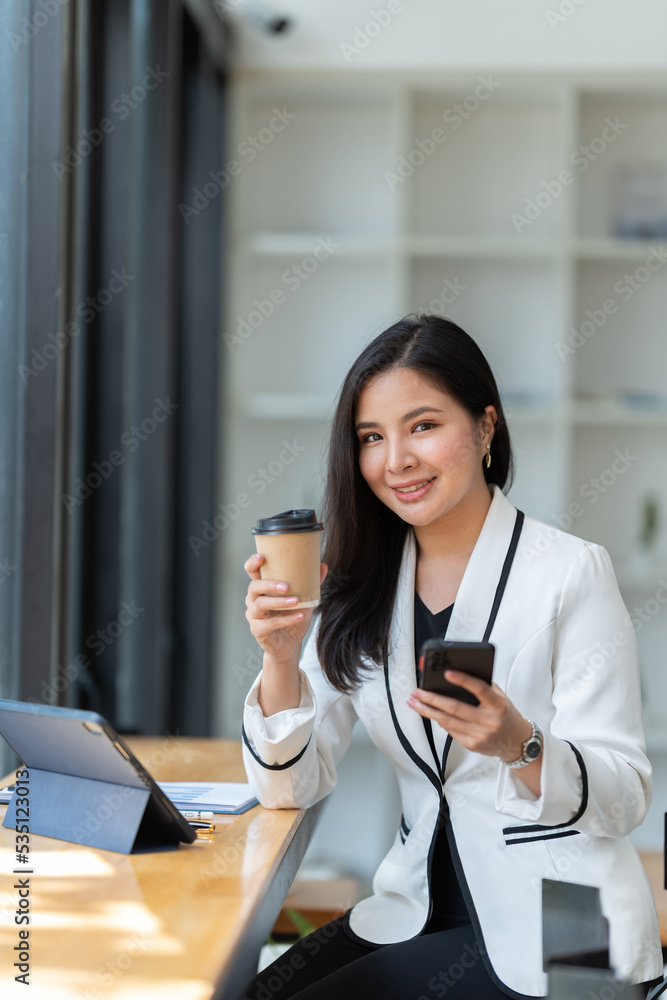 穿着正式西装的亚洲女商人在办公室里，在使用智能手机和工作时心情愉快。