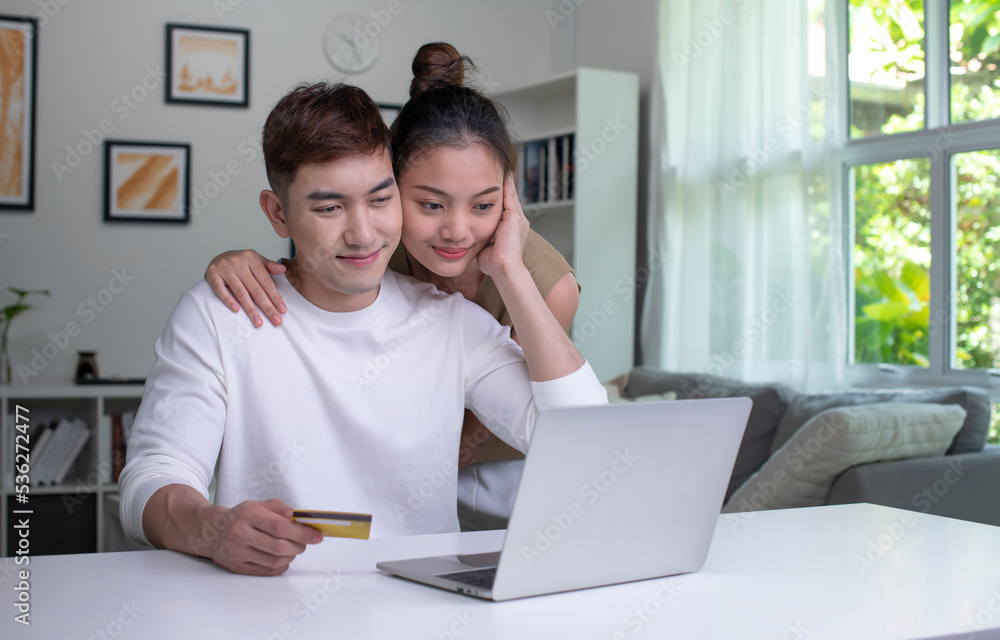 一对年轻的亚洲夫妇拿着信用卡，用着笔记本电脑。快乐的男人和女人在家工作