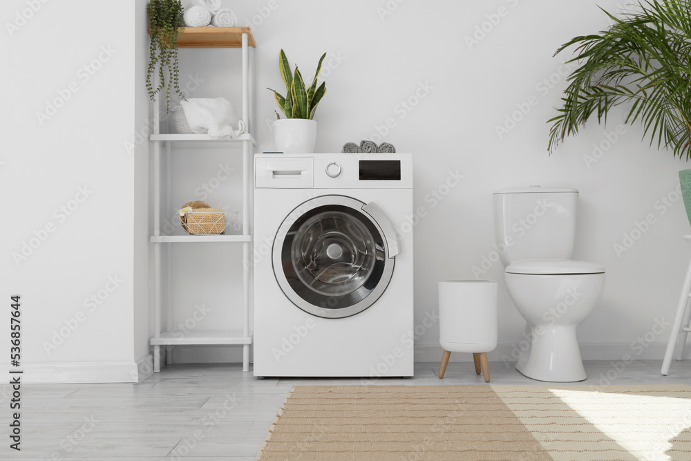 Modern washing machine, shelf unit and toilet bowl near white wall in bathroom interior