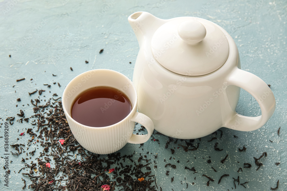 Cup of black tea, teapot and dried leaves on color background