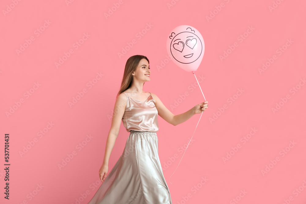 Beautiful young woman holding balloon with drawn emoticon on pink background