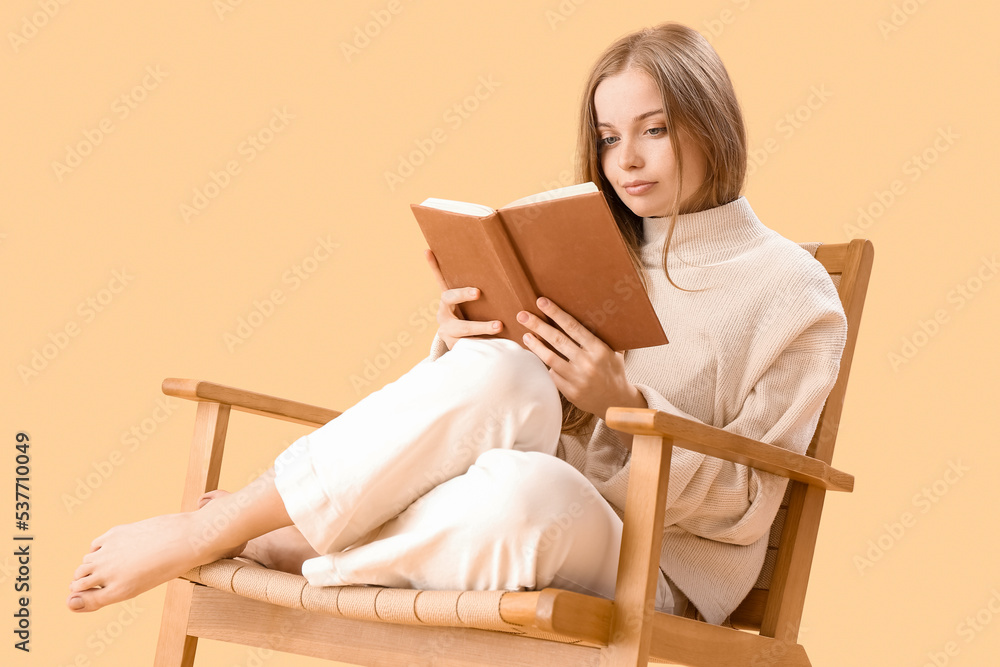 Young woman reading book in armchair on beige background