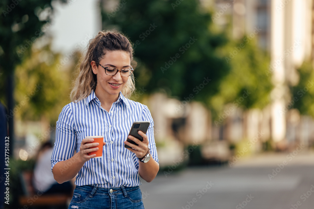 Smiling girl walking on the street, holding cup of coffee and using a phone.