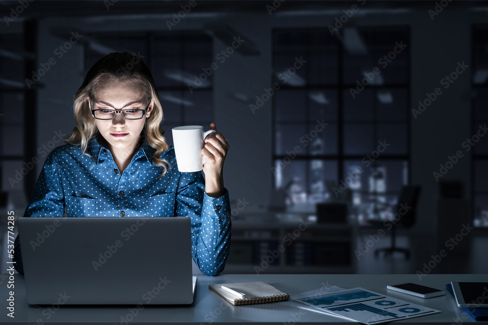 Attractive blonde working on laptop in dark office. Mixed media