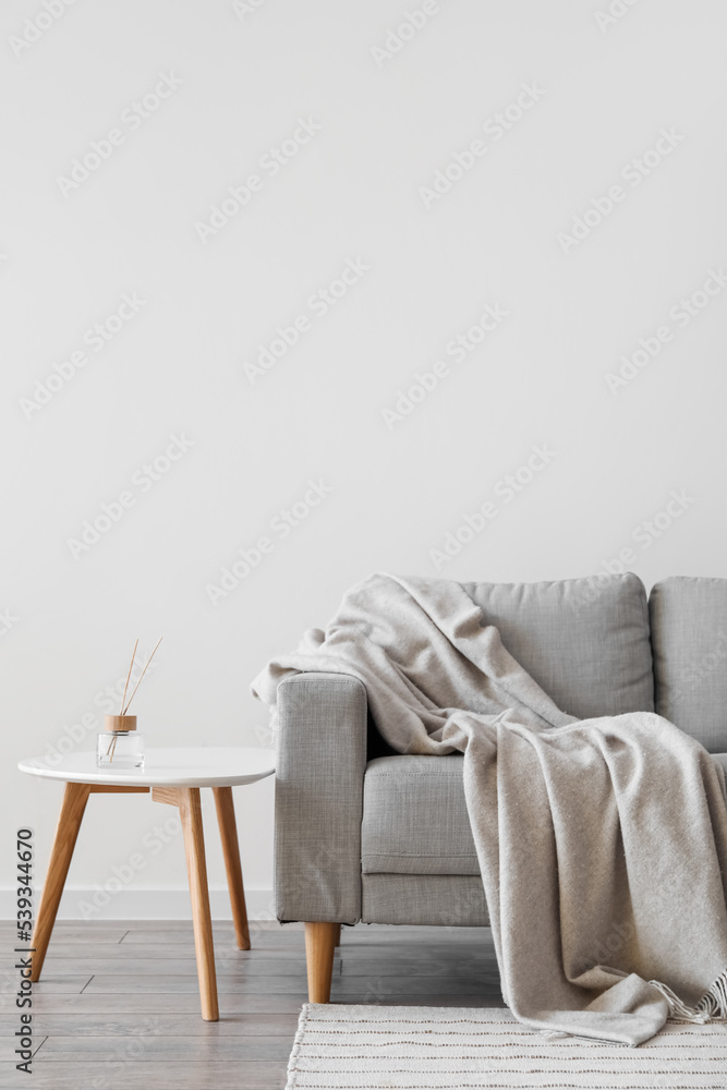 Grey sofa with plaid and reed diffuser on table near light wall