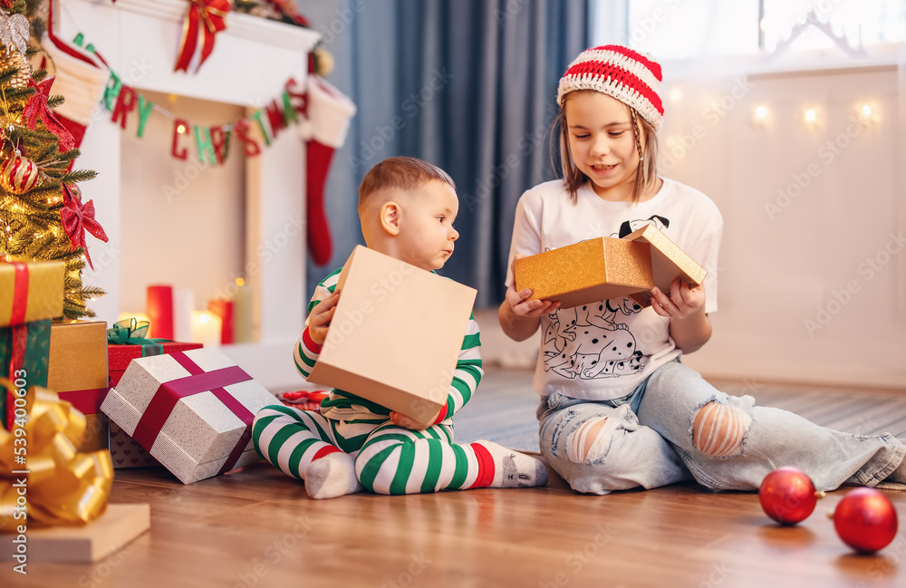 Sister and brother sitting on the floor near Christmas tree indoors.