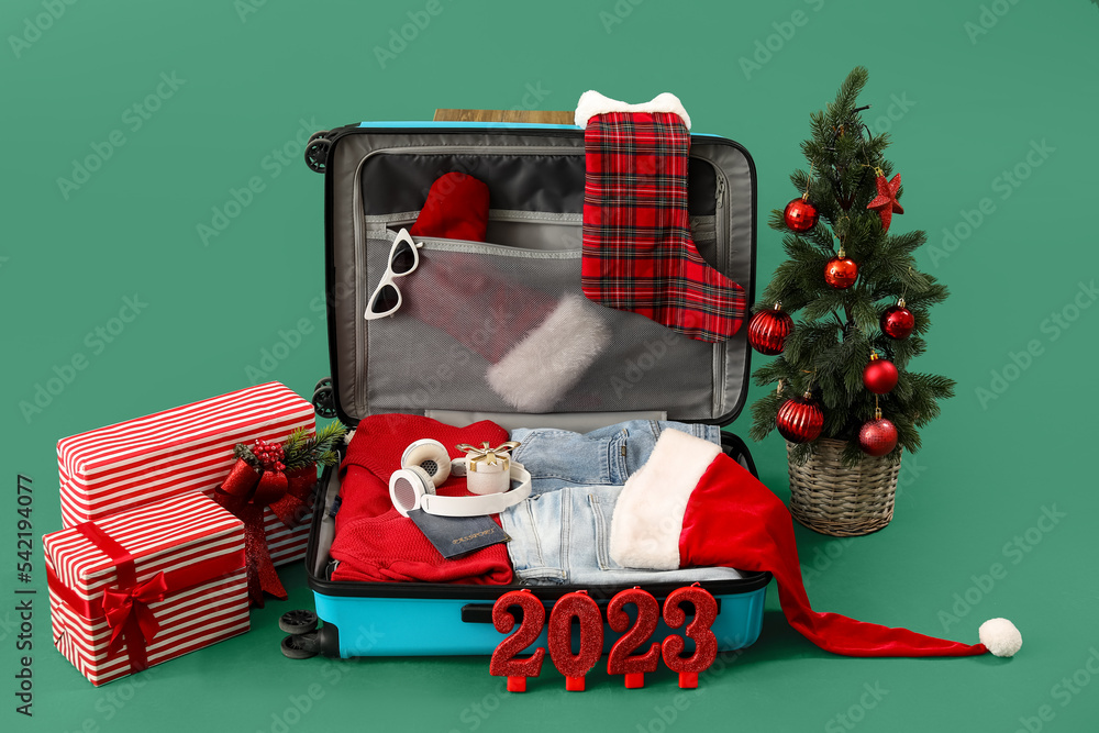 Suitcase with Christmas tree, gifts, figure 2023 and travel accessories on green background