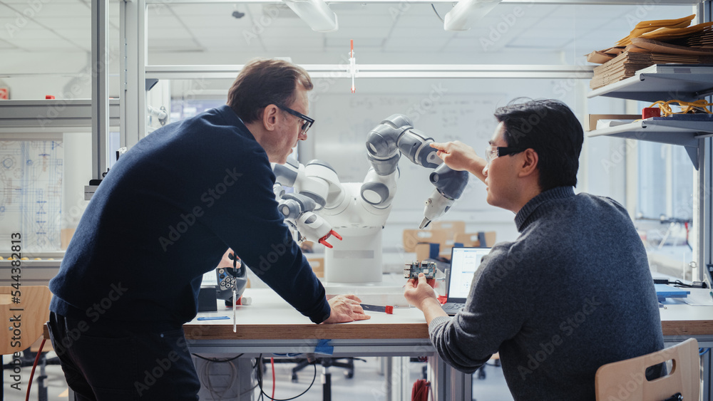 Adult Caucasian Engineer Changes Position of Robotic Arm with Controller and Having a Discussion wit