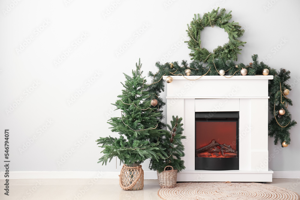 Electric fireplace with Christmas trees, wreath and fir branches near light wall