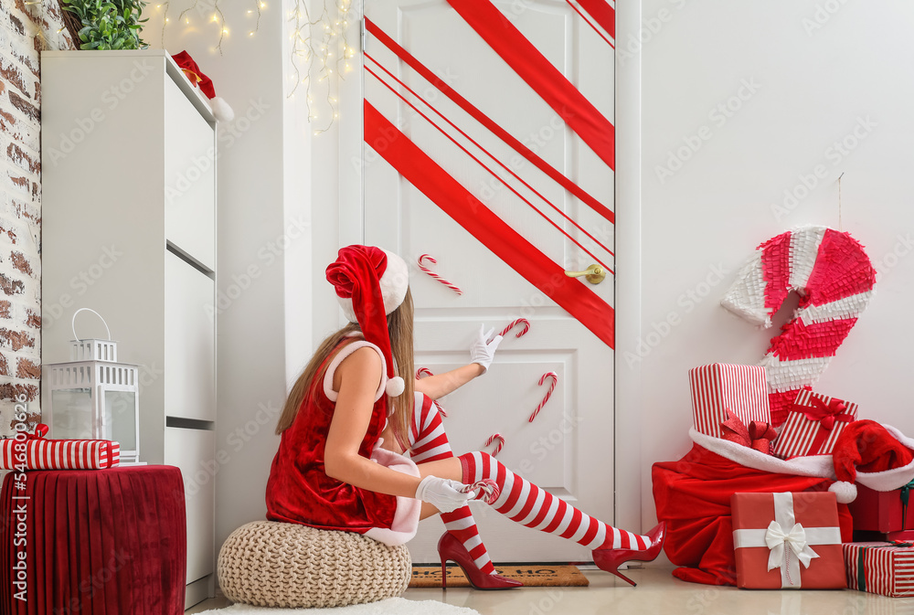 Young woman in Santa costume sitting near white door with Christmas decor