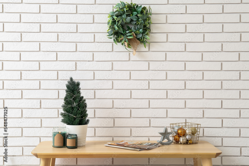 Table with fir tree, magazines, Christmas balls and mistletoe wreath on white brick wall