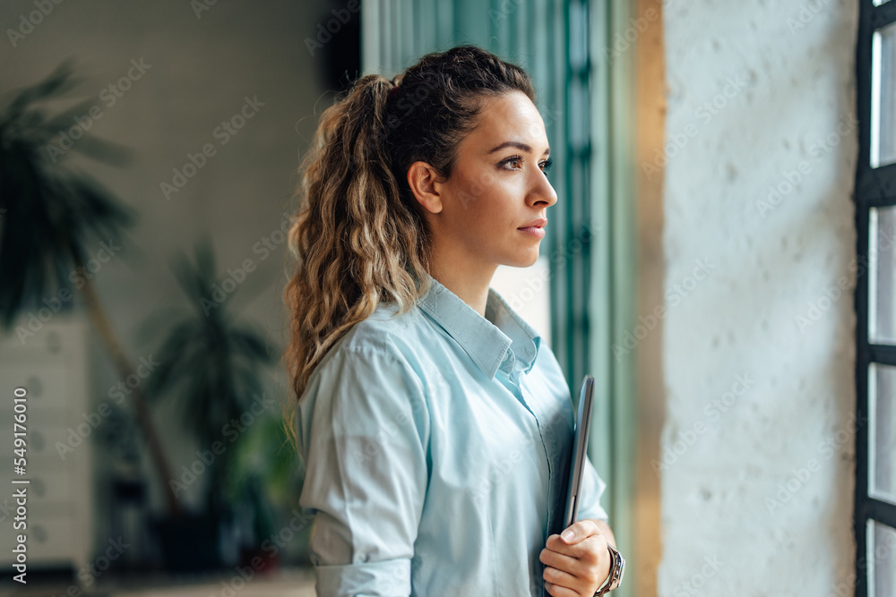 一个漂亮女孩的侧视图，她在办公室，拿着平板电脑，透过窗户看。