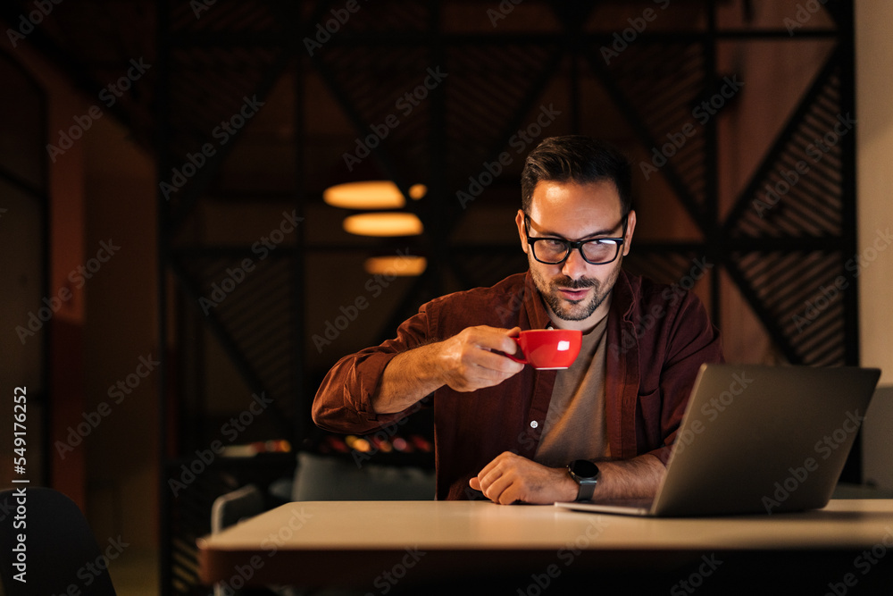 Businessman hardworking late at night, drinking coffee, using a laptop.