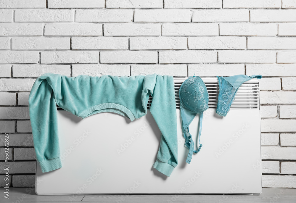 Female sweatshirt and underwear drying on electric radiator near white brick wall