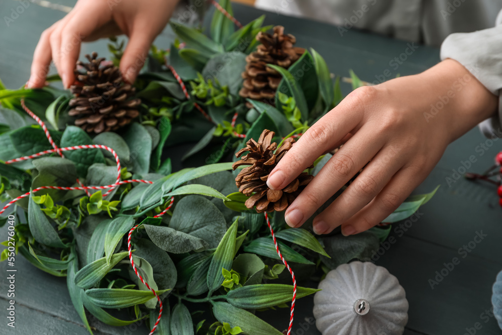 Woman decorating Christmas wreath with pine cones on color wooden background, closeup