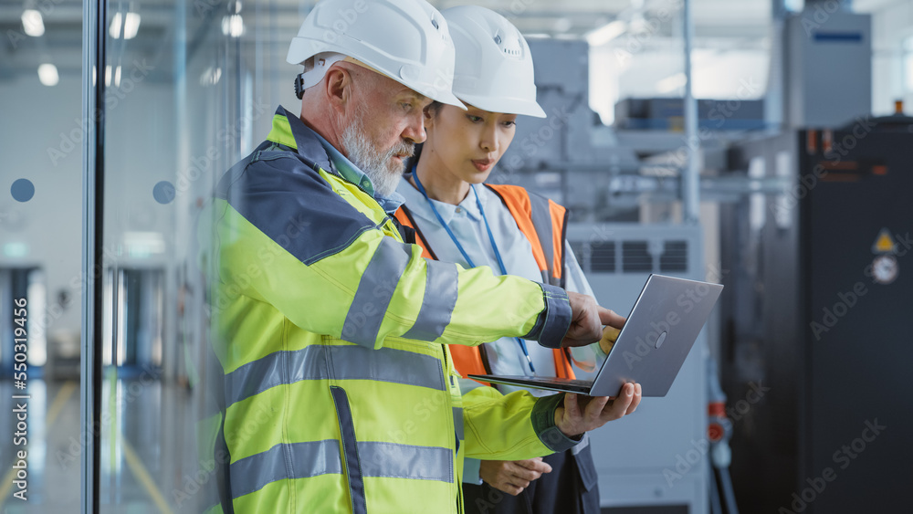 Two Diverse Heavy Industry Engineers in Safety Uniform and Hard Hats Walking with Laptop Computer an