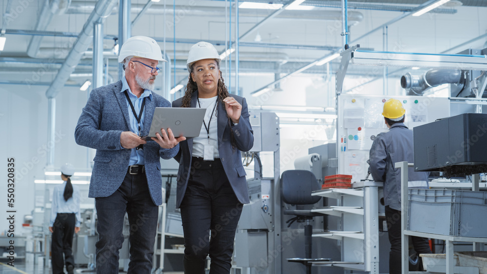 Two Heavy Industry Engineers in Hard Hats Walking with Laptop Computer and Talking in a Factory. Foo