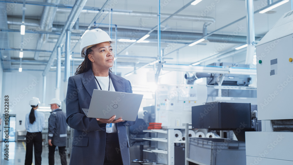 Portrait of a Black Female Engineer in Hard Hat Standing and Using Laptop Computer at Electronic Man