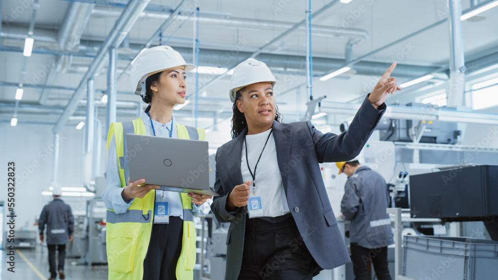 Portrait of Two Female Employees in Hard Hats at Factory. Discussing Job Assignments at Industrial M