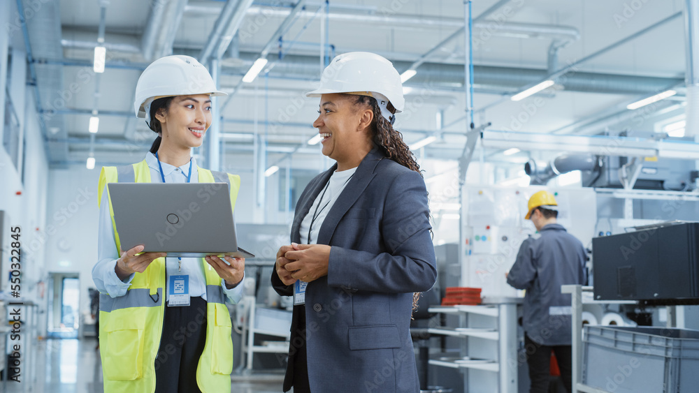 Portrait of Two Female Employees in Hard Hats at Factory Discussing Assignments at Industrial Machin