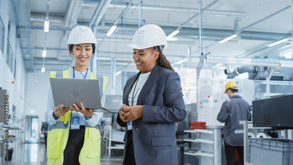 Portrait of Two Female Employees in Hard Hats at Factory Discussing Assignments at Industrial Machin