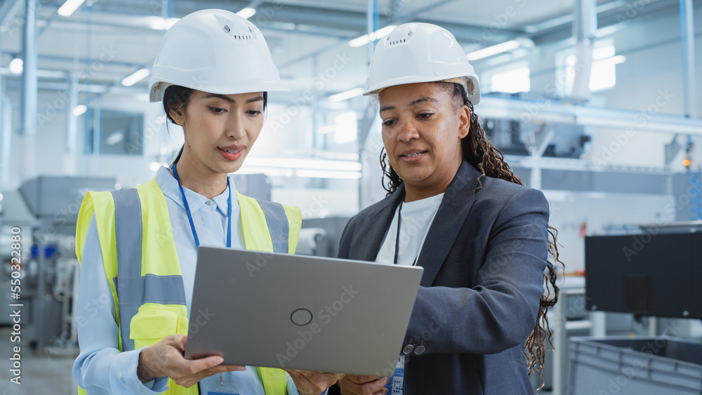 Portrait of Two Female Employees in Hard Hats at Factory Discussing Assignments at Industrial Machin