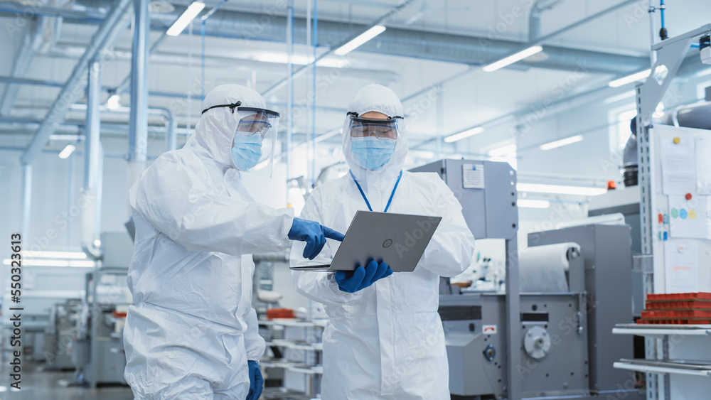 Two Scientists Walking in a Heavy Industry Factory in Sterile Coveralls and Face Masks, Using Laptop